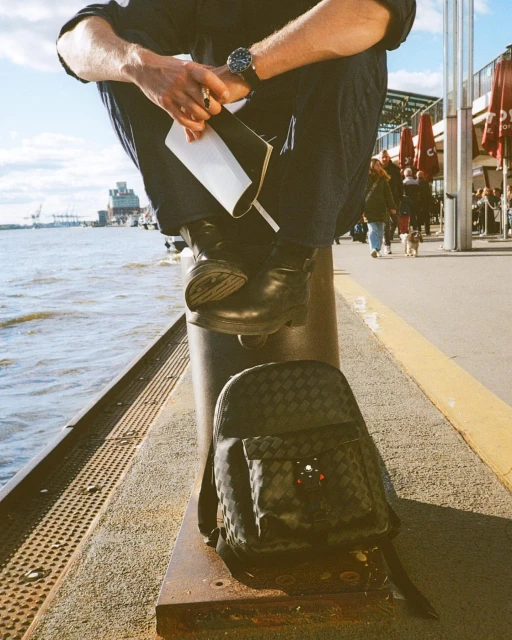 A person in a dark blue outfit sits on a bollard at a waterfront. A backpack rests against the bollard; the sea is in the background.