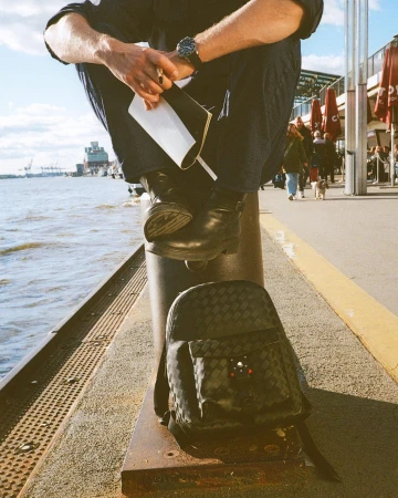 A person in a dark blue outfit sits on a bollard at a waterfront. A backpack rests against the bollard; the sea is in the background.