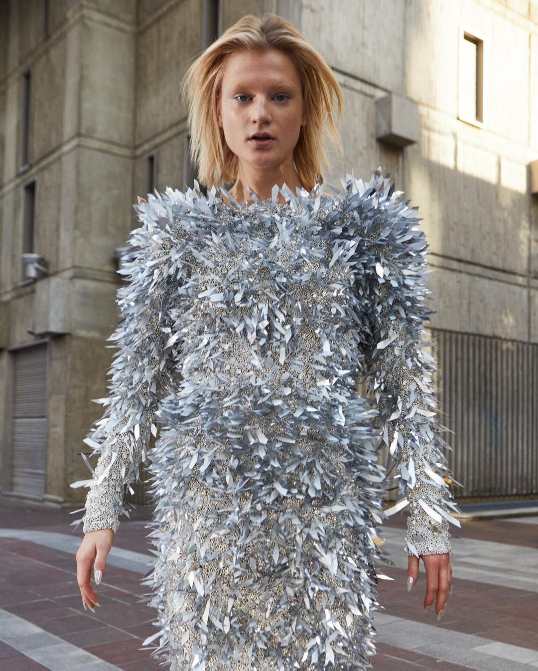 A woman in a silver textured mini dress stands outside a concrete building on a city street, posing for a fashion editorial shot.