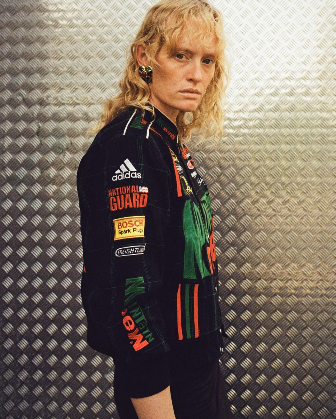 Adult female model poses in vintage-style racing jacket with brand logos against a textured metallic wall in a fashion editorial shot.