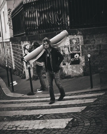 Man carrying a rolled carpet on his shoulder while walking across a city street by Julia Von Der Heide