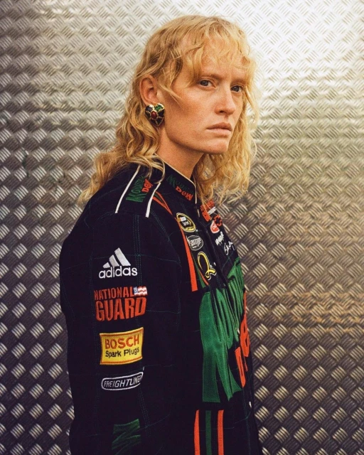 Female model with blonde curly hair wearing a racing jacket with brand logos against a metal background in a studio shot.
