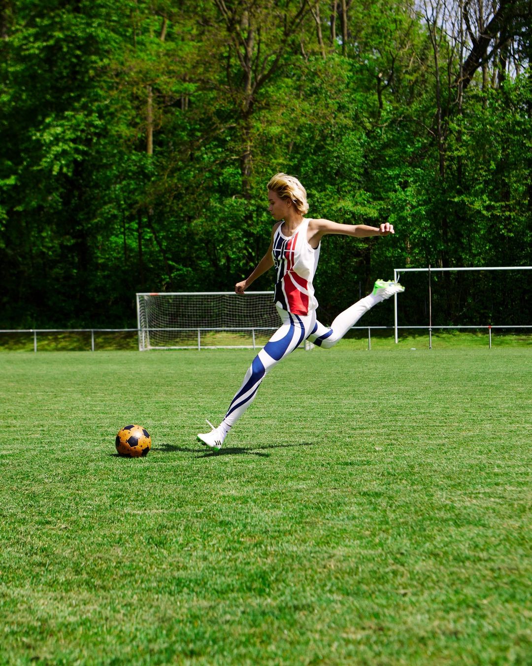 Woman in British flag sportswear kicks soccer ball on green field with trees in the background on a sunny day for sports or fitness.