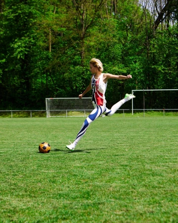 Woman in British flag sportswear kicks soccer ball on green field with trees in the background on a sunny day for sports or fitness.