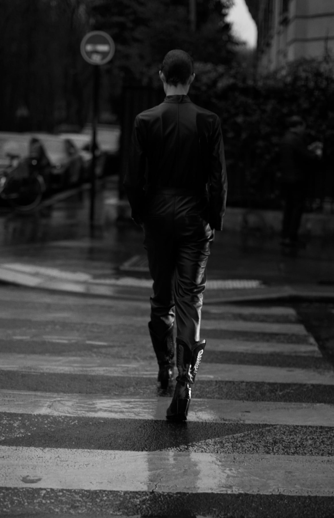 Person in dark clothing walks across a black-and-white crosswalk on a wet street; trees and building are in the background.