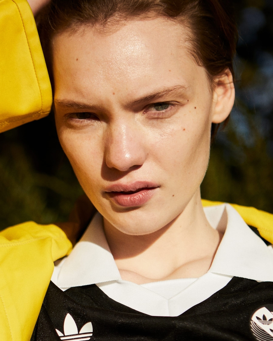 Close-up portrait of a fair-skinned adult female model with neutral expression wearing a black Adidas jersey in a natural outdoor setting.