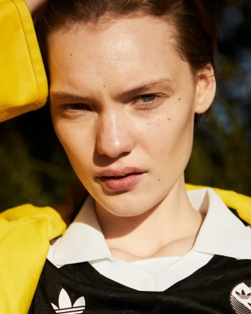 Close-up portrait of a fair-skinned adult female model with neutral expression wearing a black Adidas jersey in a natural outdoor setting.