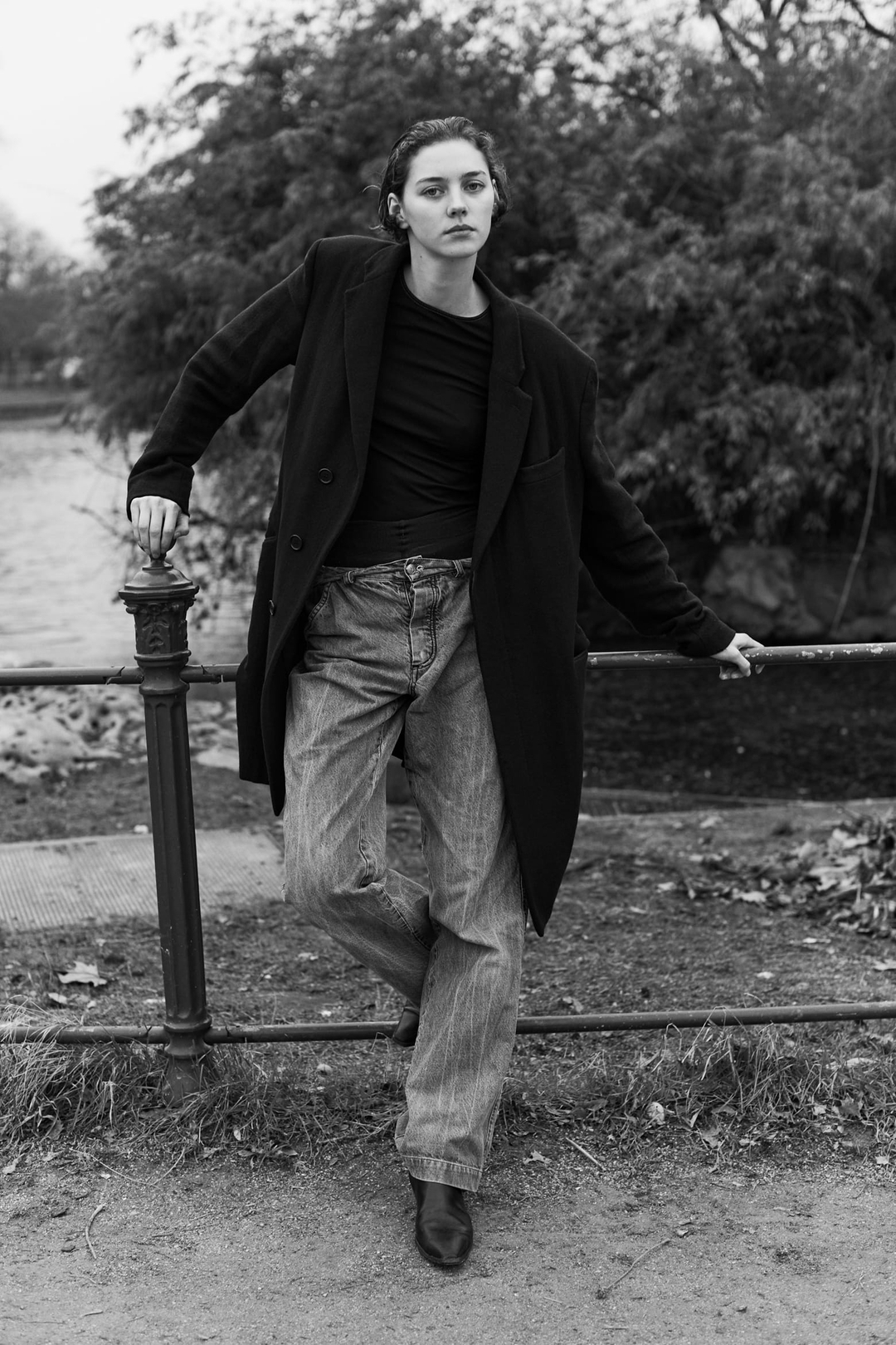 Monochrome portrait of a woman leaning against a metal railing outdoors wearing an oversized dark coat and jeans