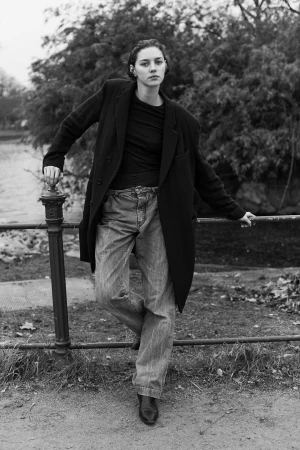 Monochrome portrait of a woman leaning against a metal railing outdoors wearing an oversized dark coat and jeans