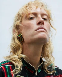 Low-angle portrait of a woman with blonde hair wearing ornate jeweled earrings and a dark patterned jacket against a white background.