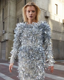 Model wearing a shimmering silver fringe dress standing outdoors in front of a concrete building in Paris.