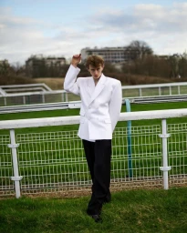 Young man in a white blazer and black trousers posing near a racetrack barrier in Paris, photographed by Julia Von Der Heide.