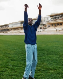 Black man in blue knit sweater and light blue trousers raising arms on a grassy sports field in Paris