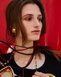 Portrait of a woman wearing beaded necklaces and gold earrings against a red backdrop, photographed by Julia Von Der Heide.