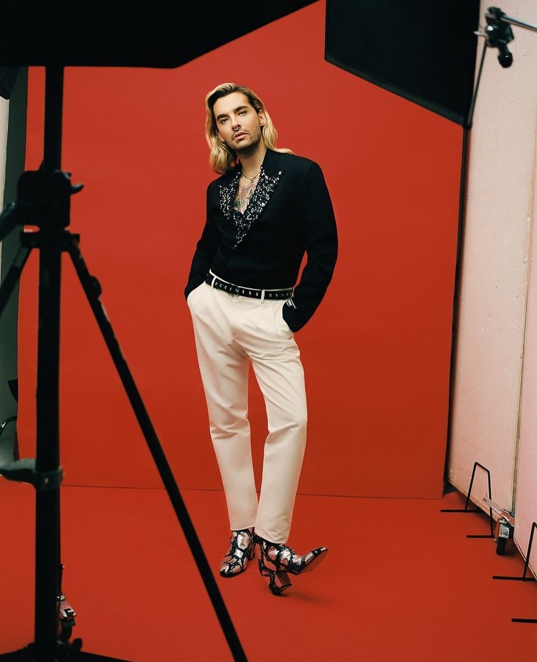 Man with long blond hair in black blazer, white pants, and snakeskin boots poses in a photo studio with a red backdrop and lighting equipment.