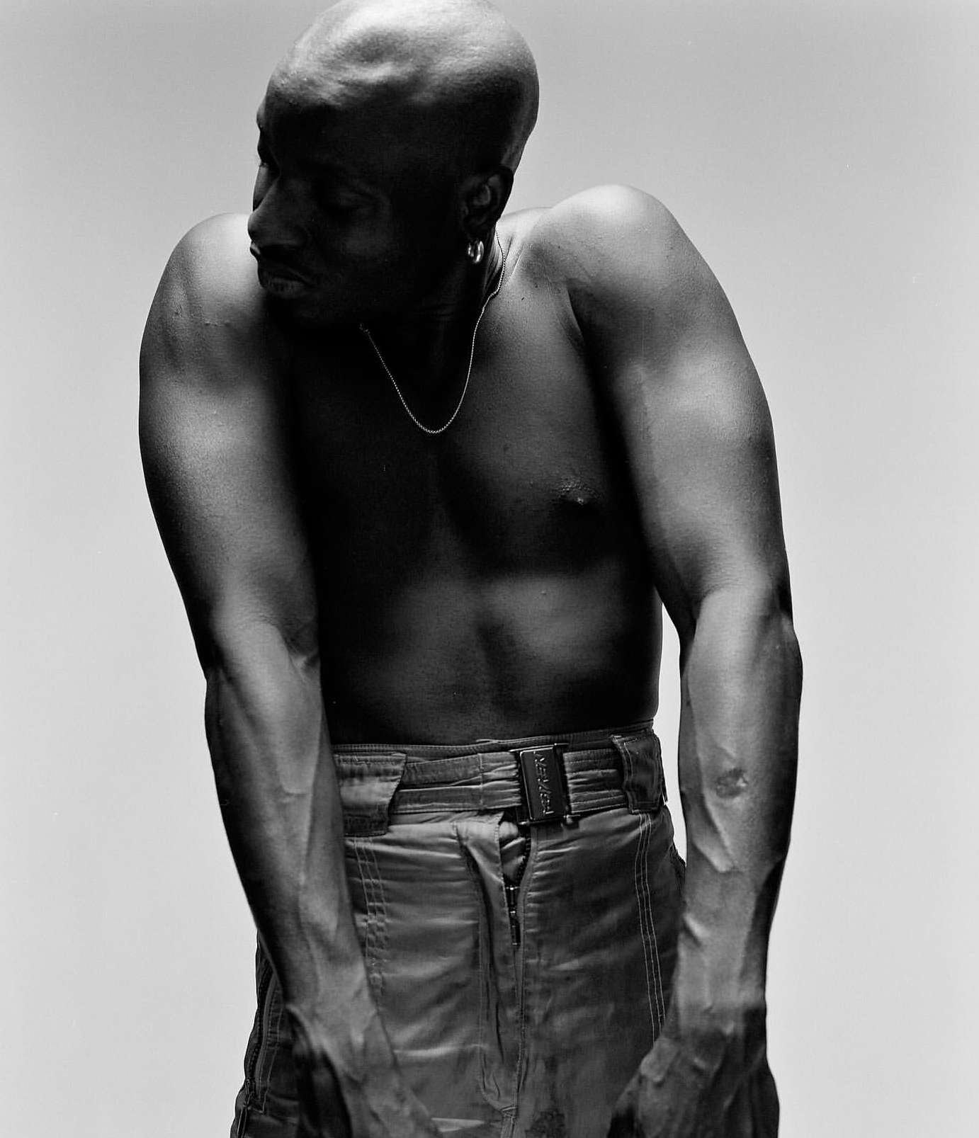 Black and white portrait of a muscular, bald, shirtless man with a silver chain and loose pants posing in a photo studio setting.