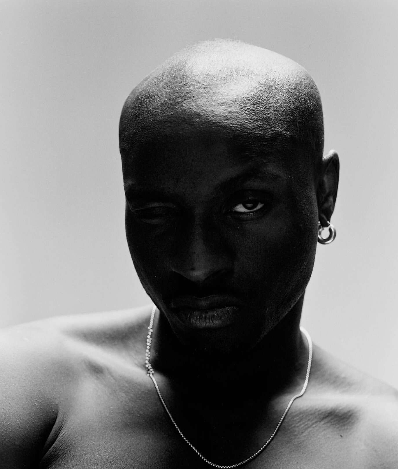 Portrait of an adult bald Black man in a close-up shot, wearing jewelry, set against a neutral background in a studio-style photograph.