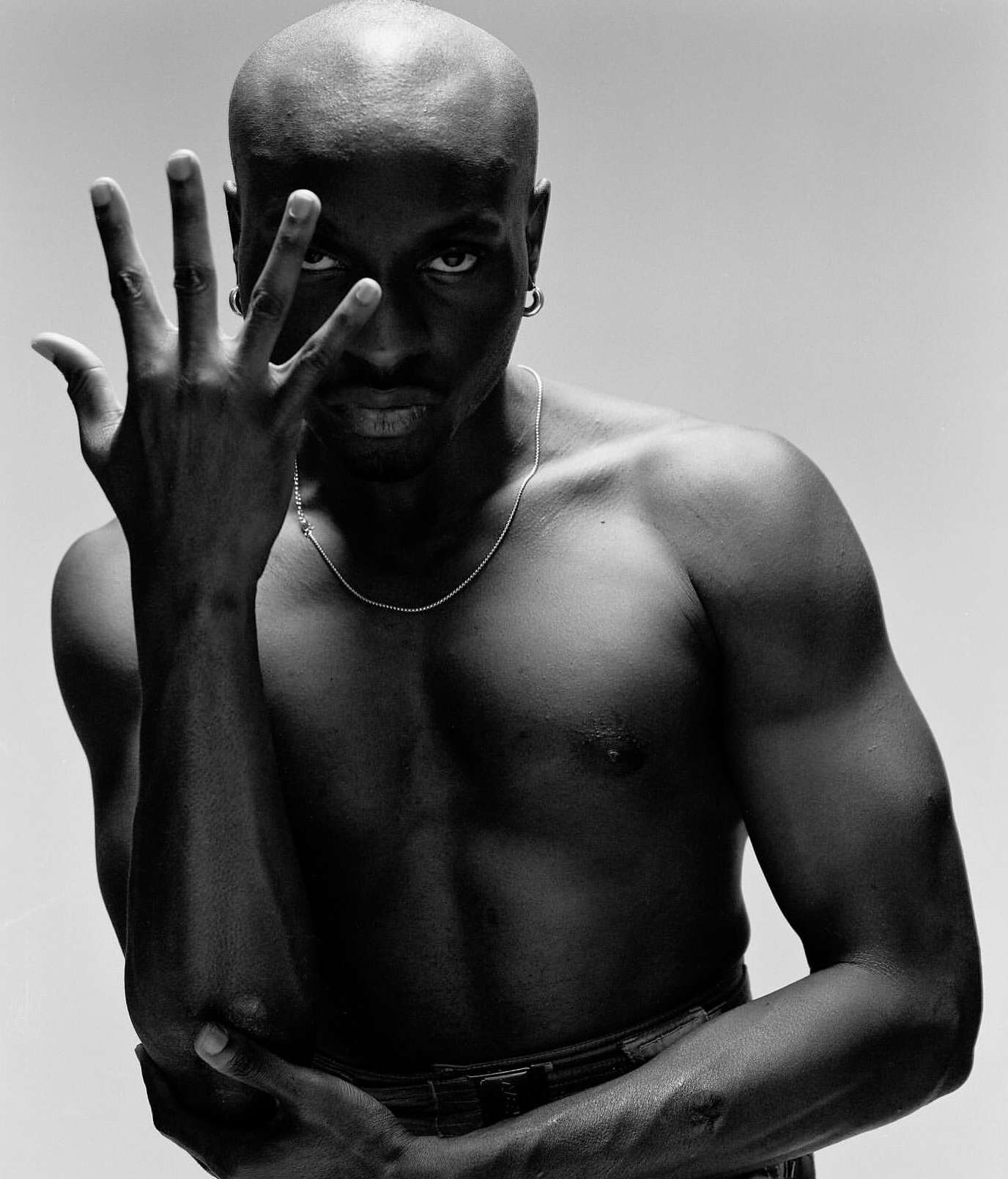 Black and white portrait of a bald, muscular, shirtless Black man with a silver chain and hand raised to his face in a photo studio.