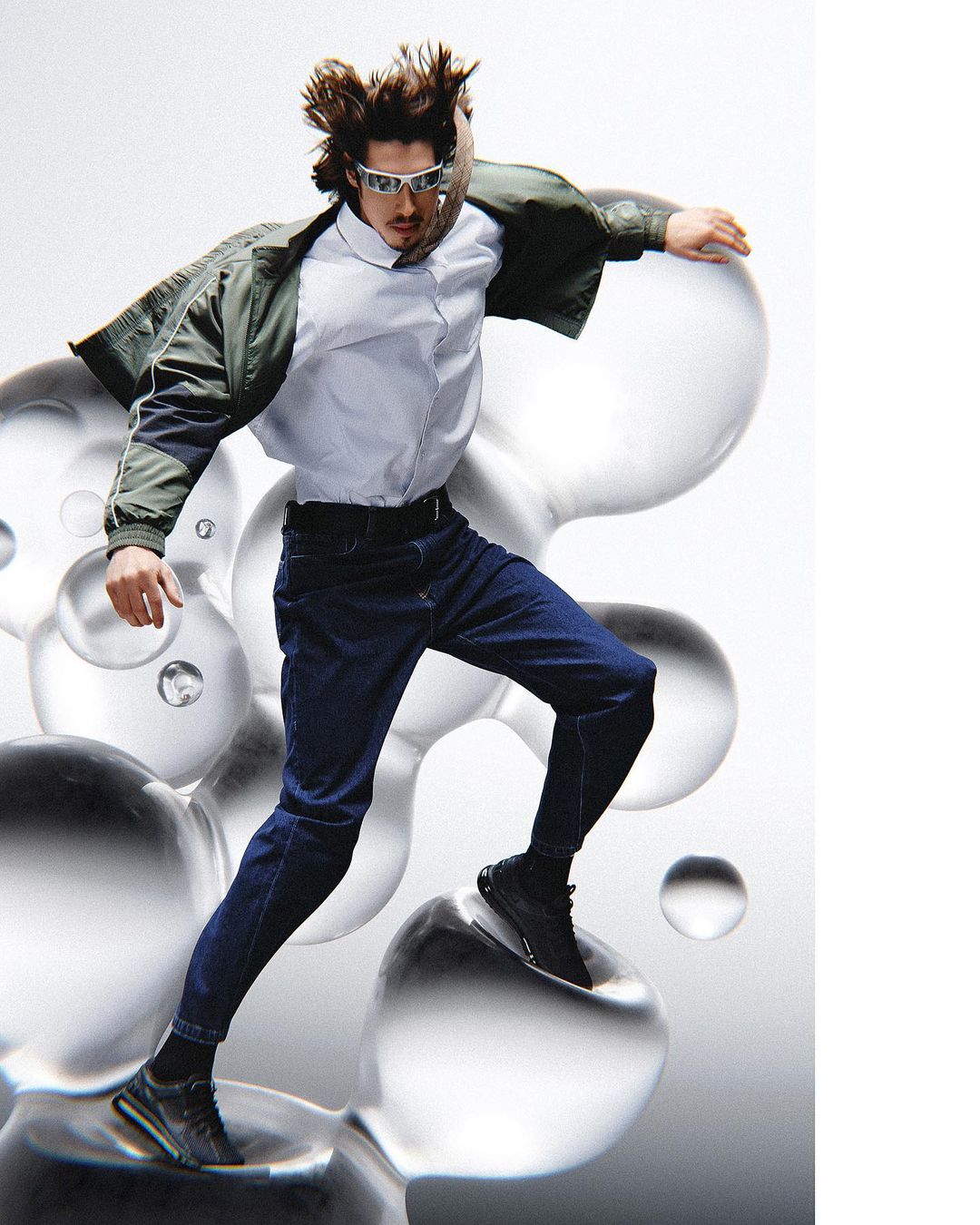 A stylish adult male model wearing a jacket, collared shirt, jeans, sunglasses, and black sneakers poses dynamically against a white background with transparent spheres.