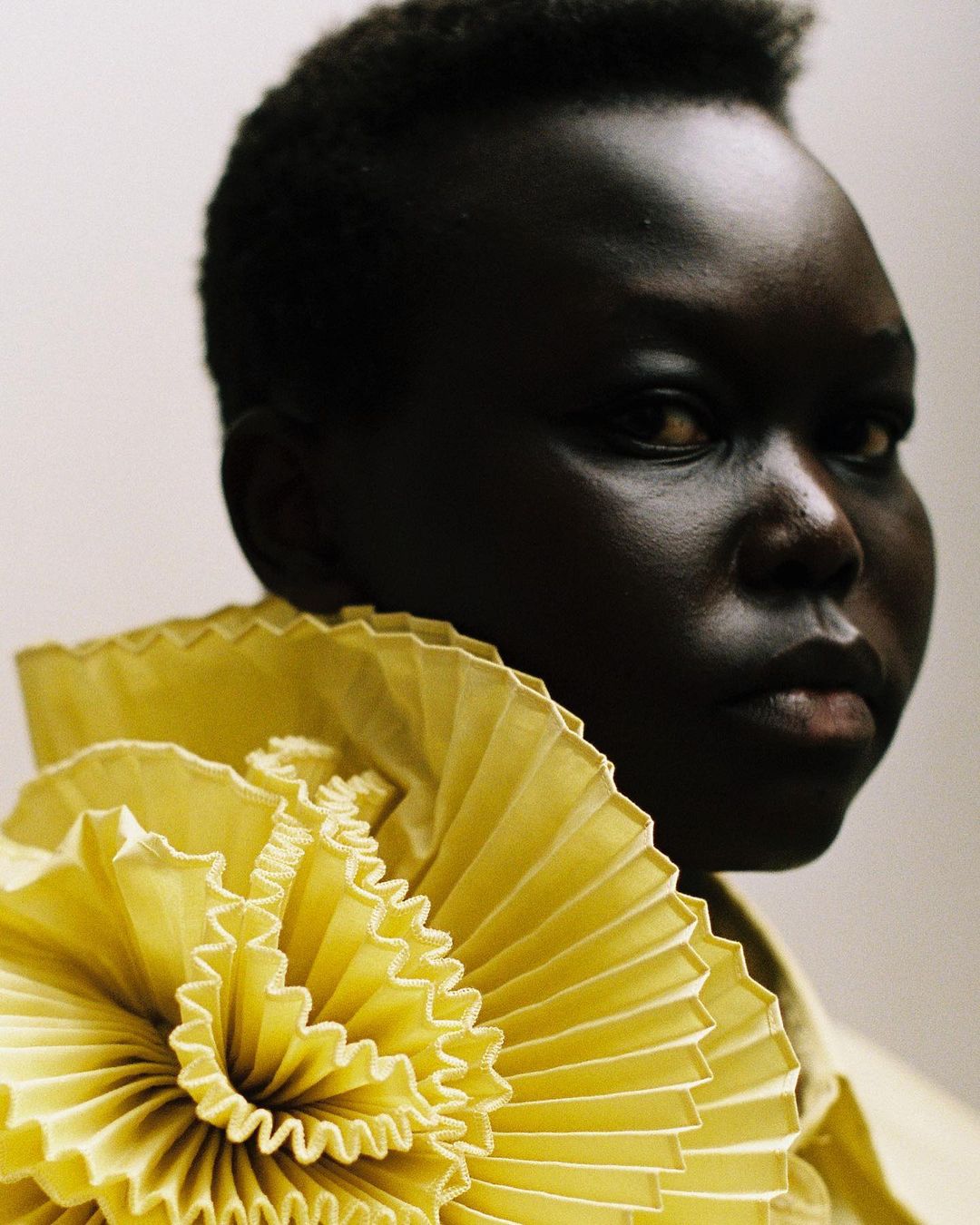 Close-up portrait of a Black woman with short hair wearing a yellow ruffled collar against a neutral backdrop, beauty and fashion focused.