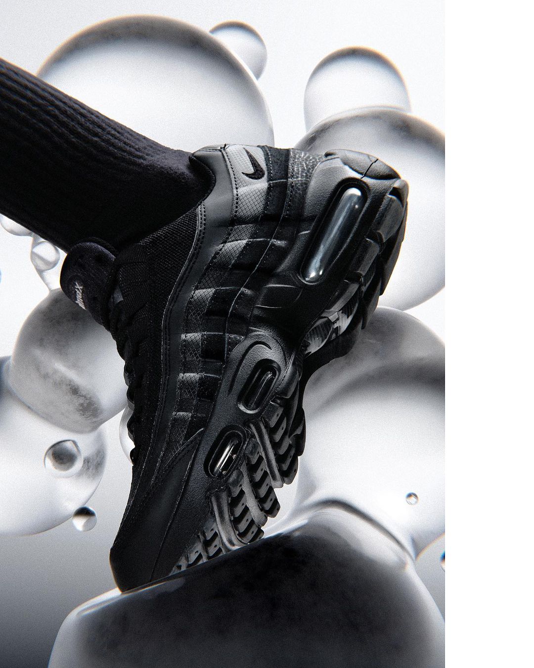 Black Nike Air Max 95 sneaker with a black sock on a white backdrop with translucent spheres, lit with bright studio lighting.