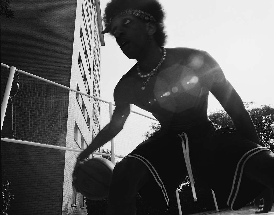 Monochromatic shot of a young Black man dribbling a basketball on an outdoor court, wearing athletic shorts and jewelry against a brick building.
