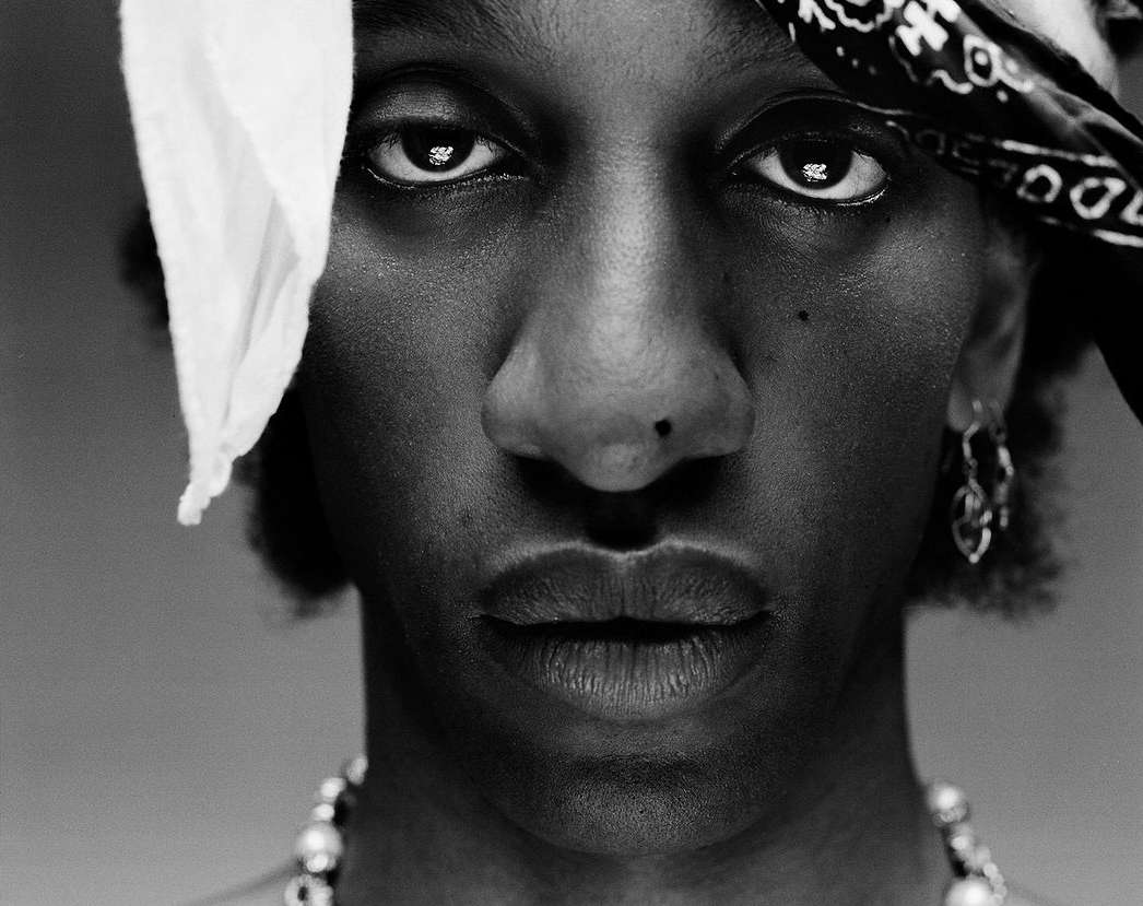 Close-up black and white portrait of a Black adult male wearing a white headscarf, beaded necklace, and hoop earrings, against a grey backdrop.