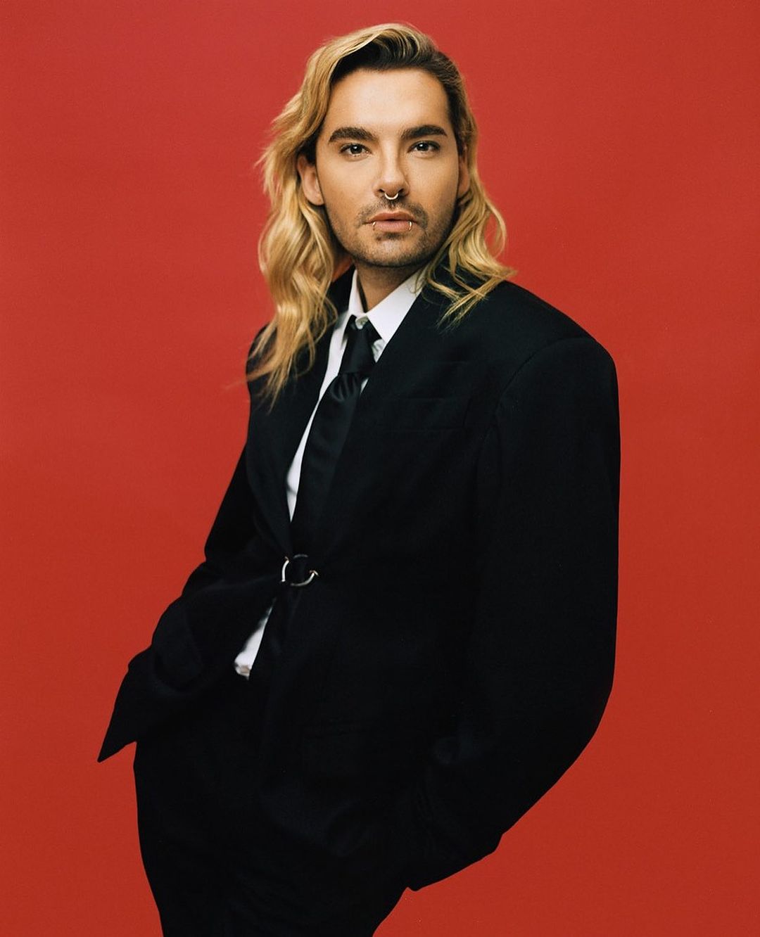 Adult male with long blond hair in black suit and tie poses against a red background in a studio portrait for fashion or editorial use.