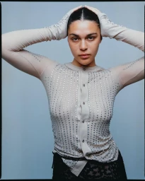 Model with dark hair wearing a crystal-embellished beige top with hands raised to head against a plain grey background