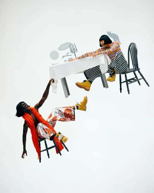 Two fashion models floating on chairs at a table, wearing colorful outfits and boots, against a white background in a studio setting.