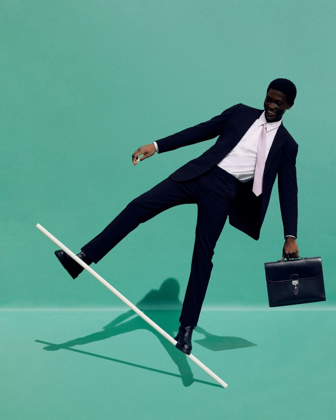 An African American businessman balancing on a white bar with a briefcase in hand against a green background in a studio setting.