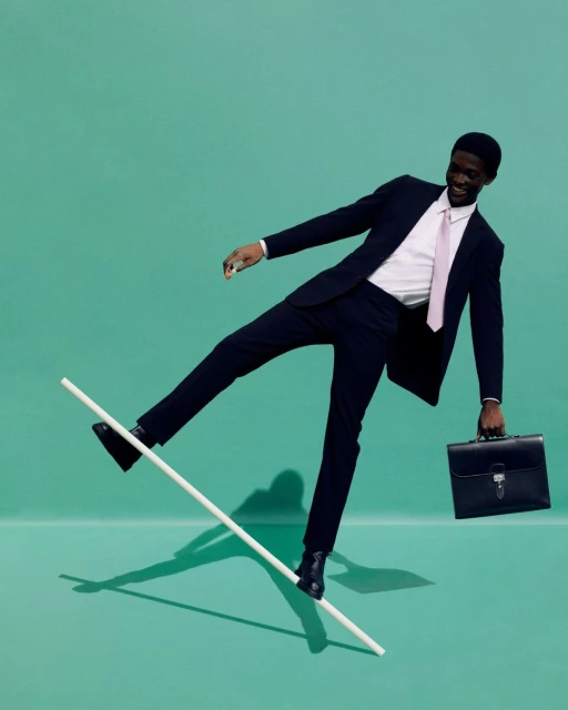 An African American businessman balancing on a white bar with a briefcase in hand against a green background in a studio setting.
