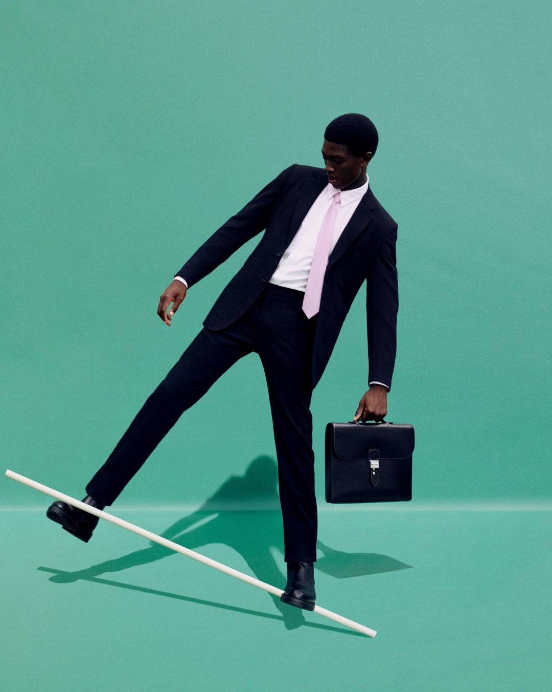 Man in a suit balancing on a pole and holding a briefcase against a teal background in a studio environment for a fashion editorial.