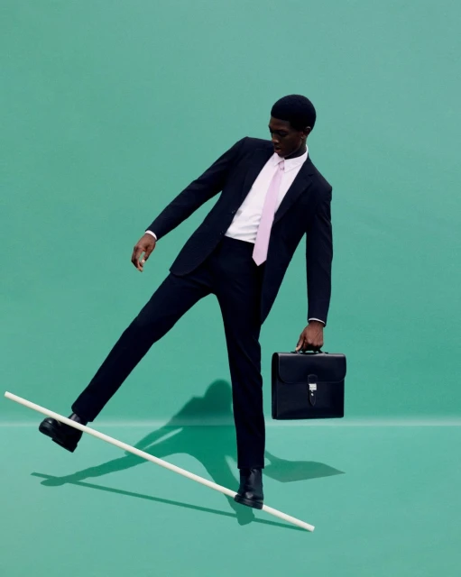 Man in a suit balancing on a pole and holding a briefcase against a teal background in a studio environment for a fashion editorial.