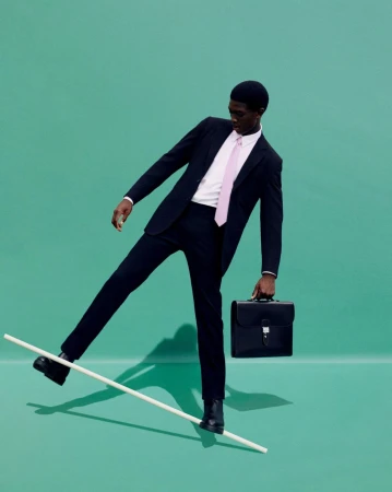 Man in a suit balancing on a pole and holding a briefcase against a teal background in a studio environment for a fashion editorial.