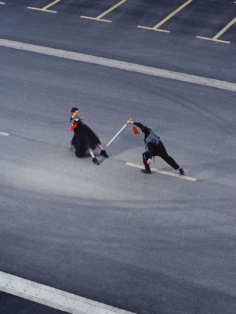 Two people in an outdoor asphalt parking lot holding a pole, dressed in black outfits. Woman in a black tutu-style skirt.