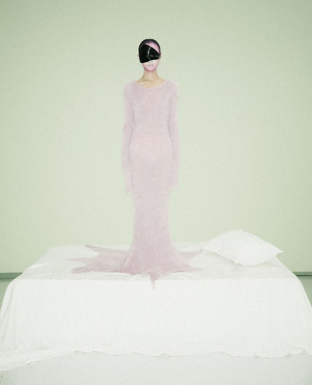 A woman stands on a bed wearing a long pink dress and a large, black eye covering in a surreal, fine art fashion photography style.