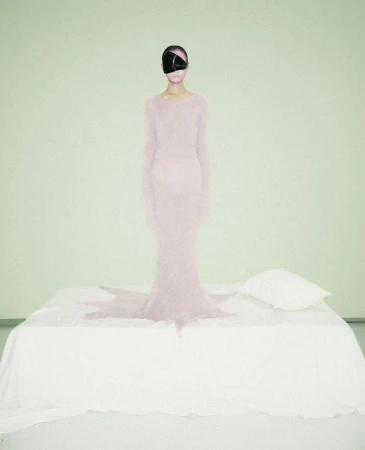 A woman stands on a bed wearing a long pink dress and a large, black eye covering in a surreal, fine art fashion photography style.