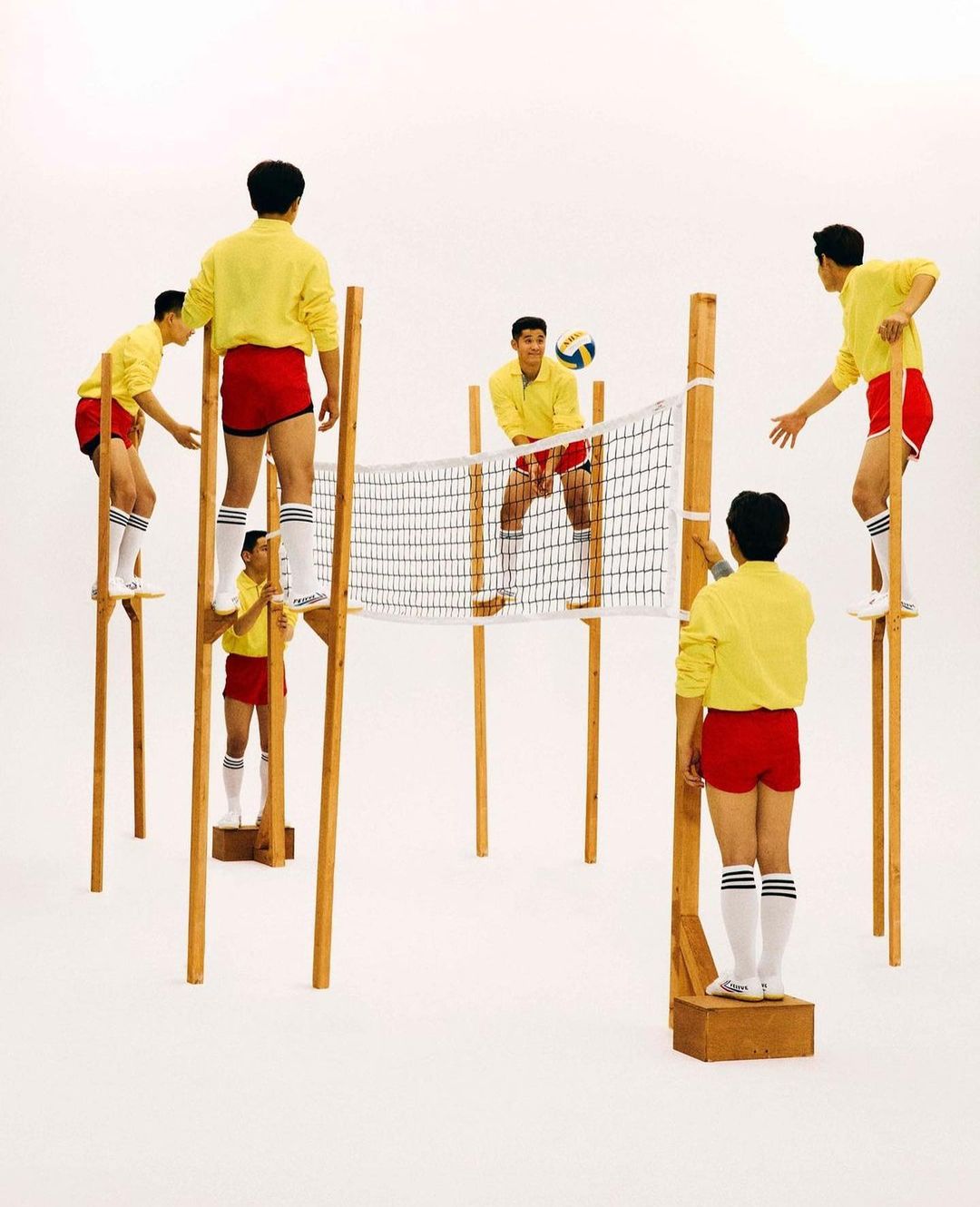 Several young men in yellow shirts and red shorts playing volleyball on stilts on a white background, creating a playful, dynamic composition.