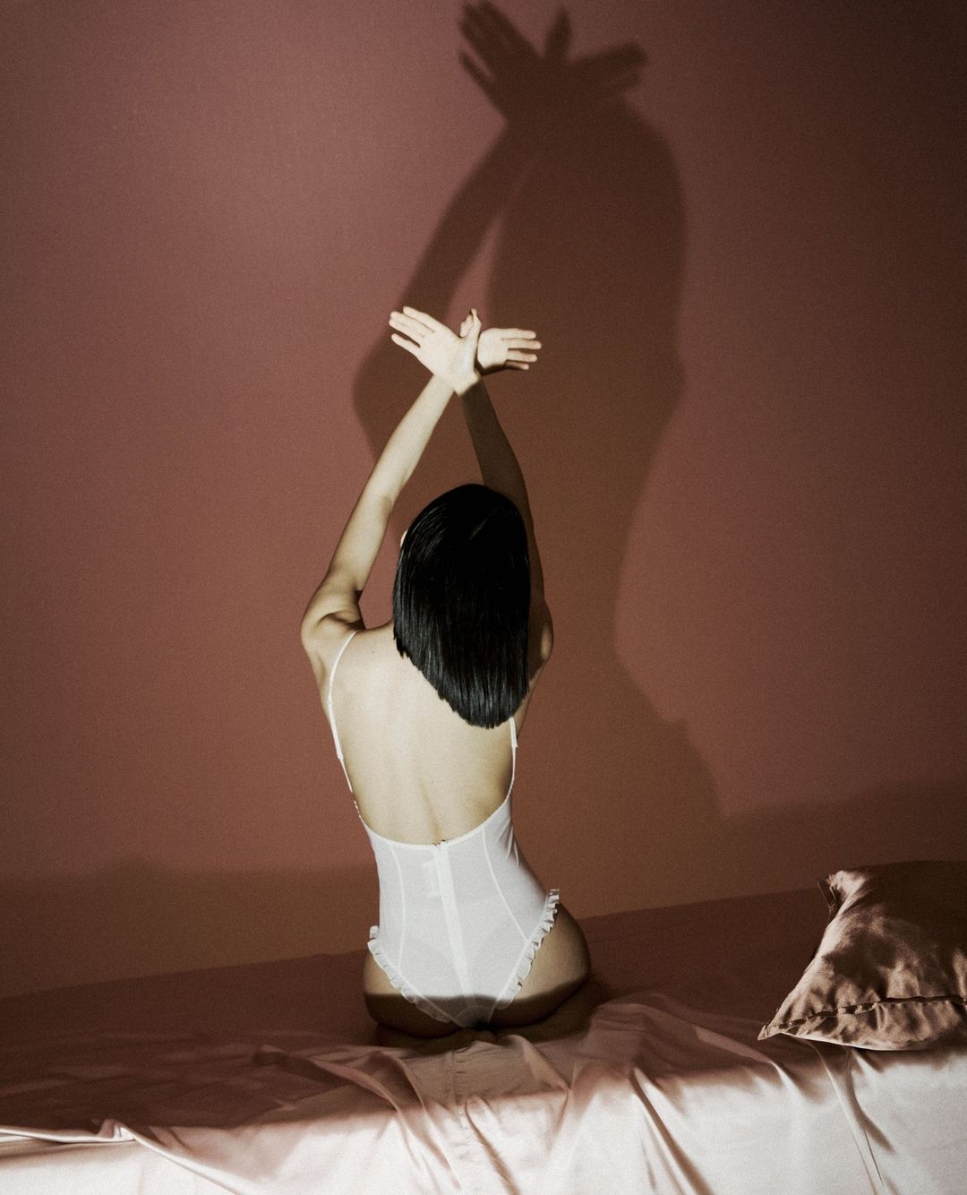 Back view of a person in white lingerie sitting on a bed, arms raised to cast a shadow on the wall.