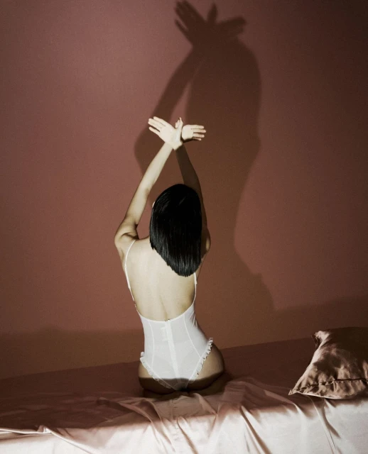 Back view of a person in white lingerie sitting on a bed, arms raised to cast a shadow on the wall.