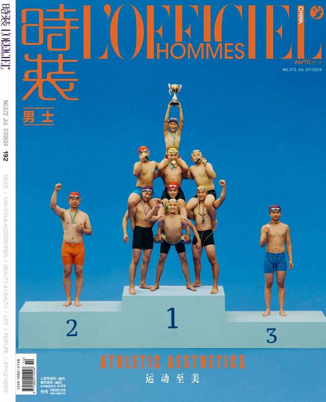 Men's fashion magazine cover featuring a pyramid of seven men wearing swimwear and medals, one holding a trophy on a blue background.