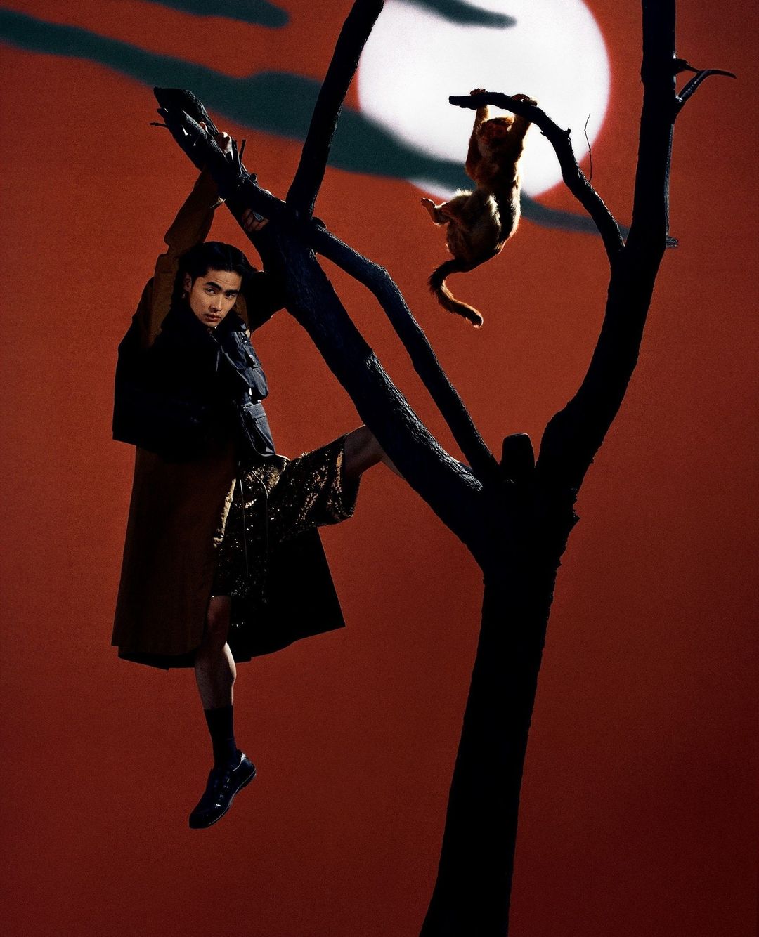 Stylish adult male poses in brown coat and shorts next to bare tree with hanging monkey figure, set against an orange background with a moon.