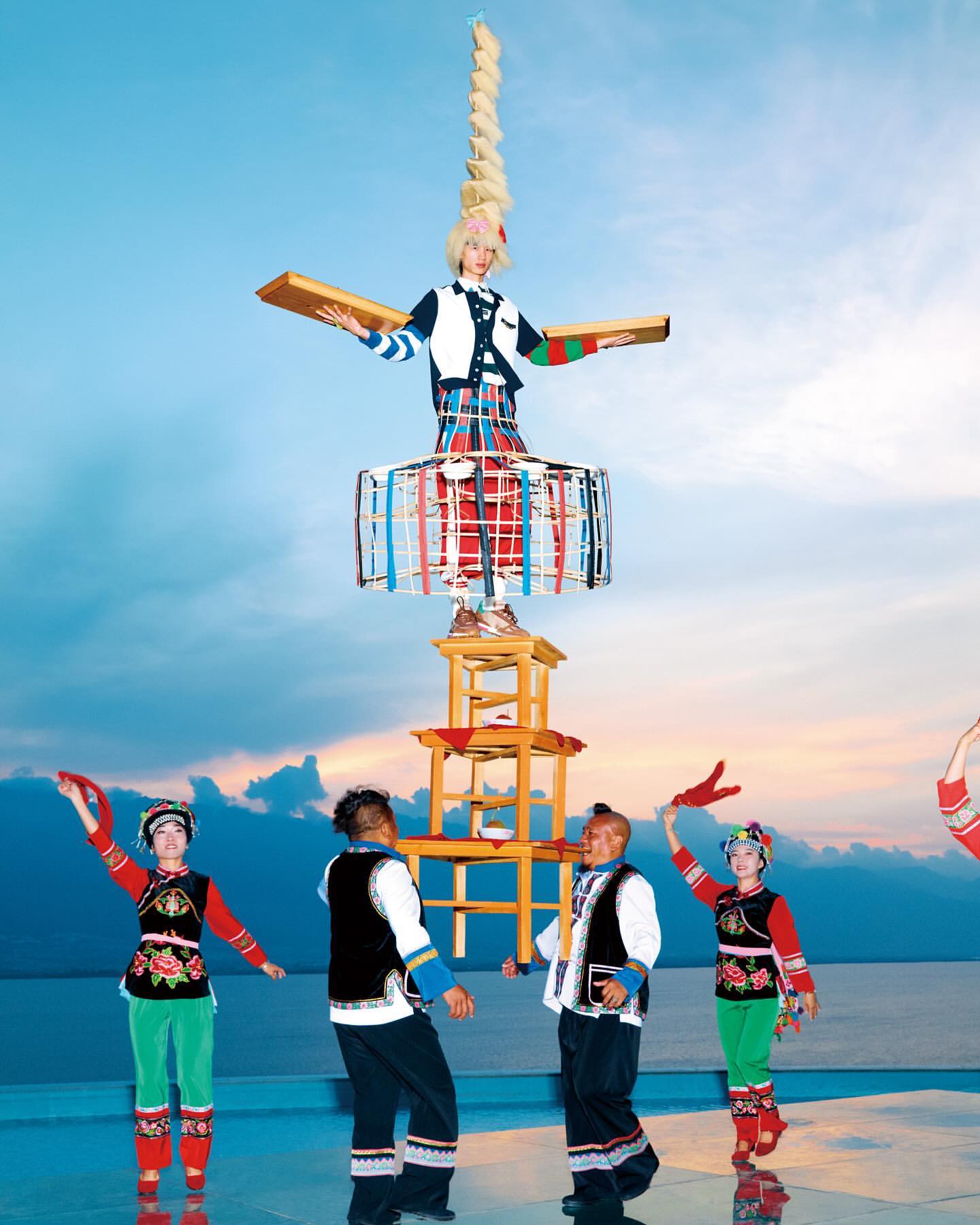 A woman balances atop a tall structure of chairs and a cylindrical form, surrounded by several people in a bright outdoor setting.