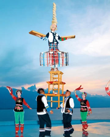 A woman balances atop a tall structure of chairs and a cylindrical form, surrounded by several people in a bright outdoor setting.