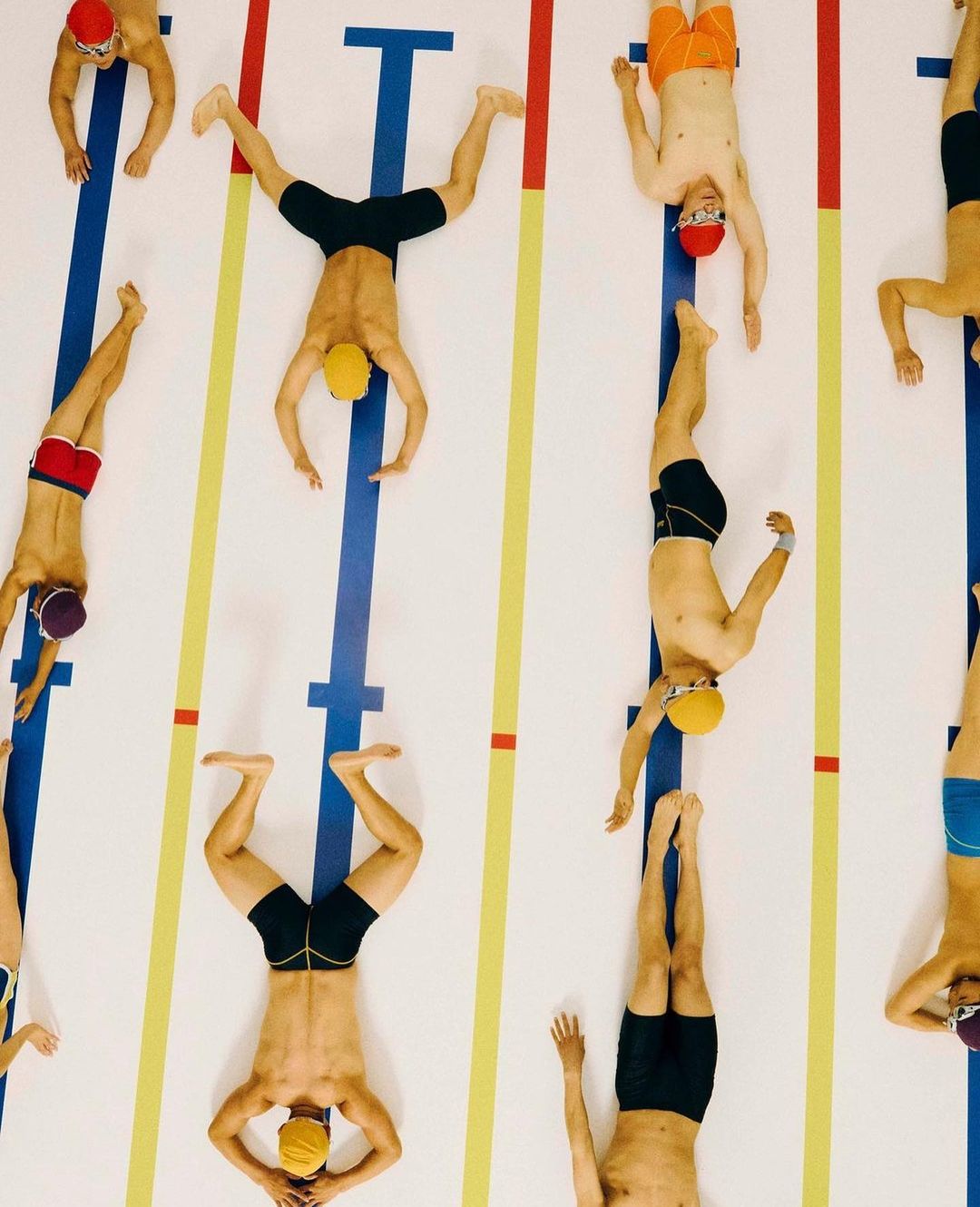 Overhead shot of multiple swimmers laying head-to-toe along the pool lane lines wearing swimming caps and trunks on the pool deck.