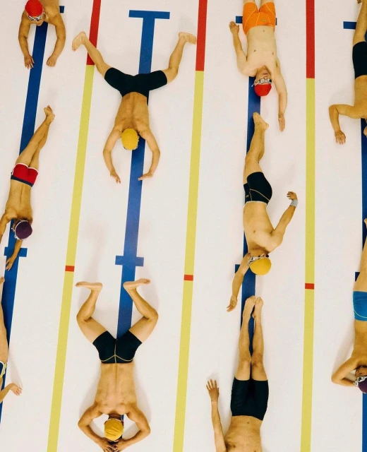 Overhead shot of multiple swimmers laying head-to-toe along the pool lane lines wearing swimming caps and trunks on the pool deck.