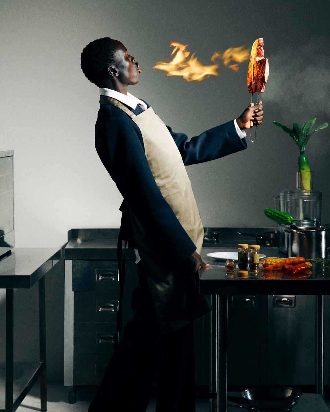 An adult male blowing fire over food held on a metal skewer in a commercial kitchen with metal counters and stainless steel appliances.