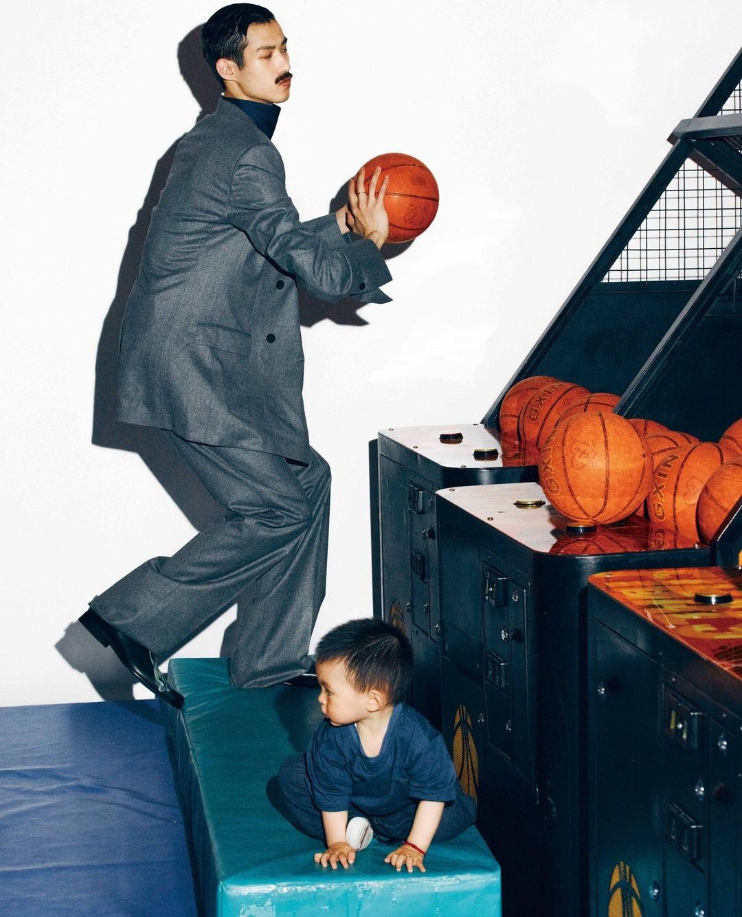 Adult male in a grey suit with a basketball next to a child sitting near a basketball arcade game machine against a white background.