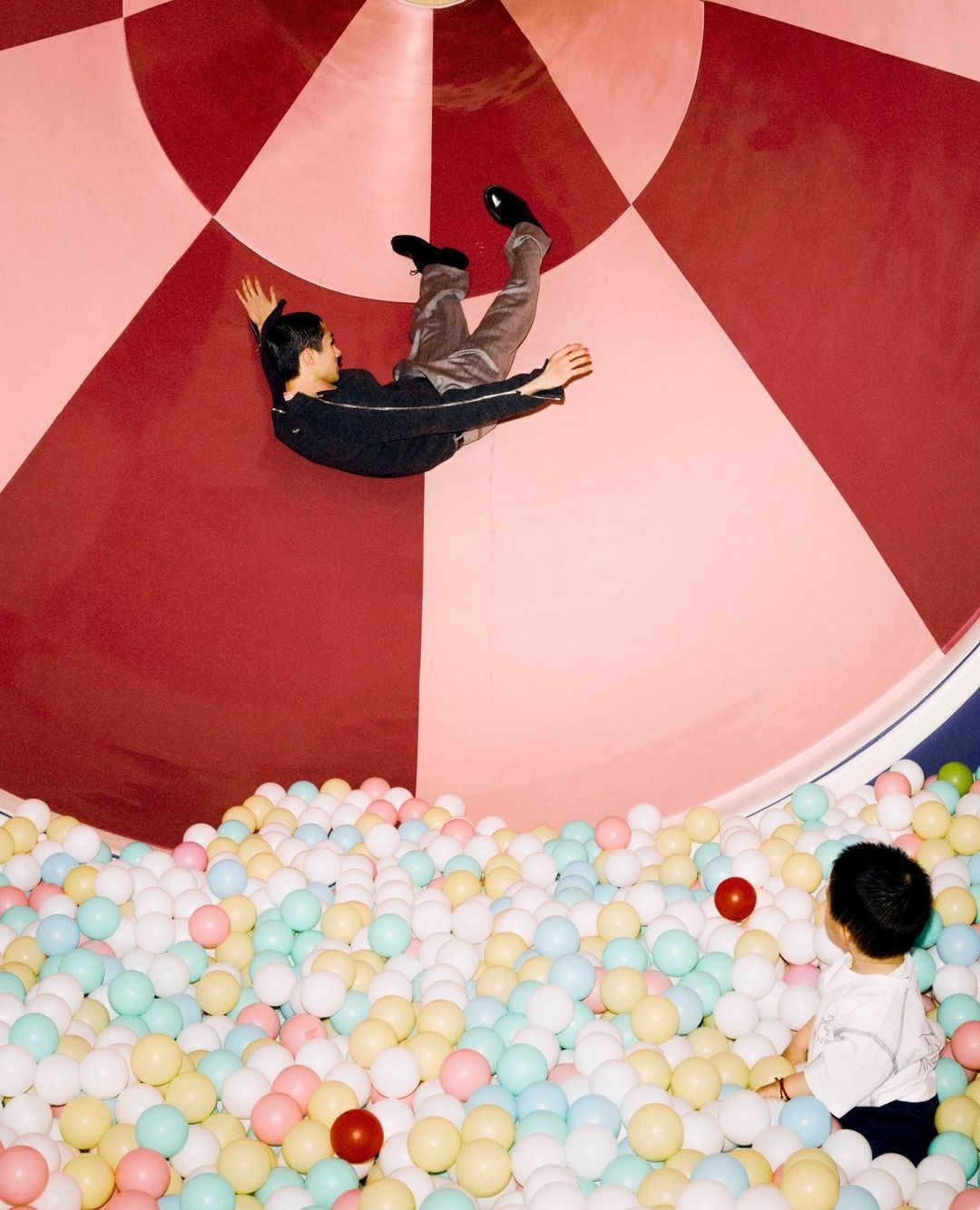 Adult and child play in a pink-and-red circular pit filled with colorful balls, creating a whimsical and playful scene indoors.